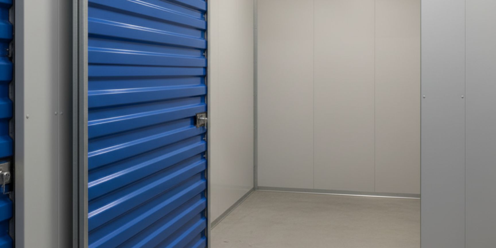 Close-up of a single, lockable self-storage compartment with a blue roller door. The door is slightly open, revealing the empty, clean storage room, ready for storing personal belongings or smaller business goods. The wall of the storage box is a neutral shade of grey, which emphasises the practicality and protection of the contents.